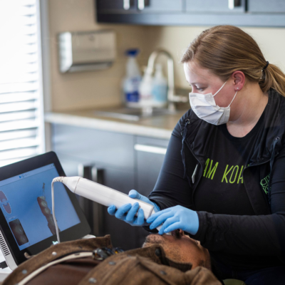 A Kohrs Orthodontics team member wearing a face mask and blue gloves uses a handheld 3D oral scanner to take digital impressions of a patient’s teeth while viewing the digital image on a nearby computer monitor in a modern dental office, representing how digital impressions transform your orthodontic experience