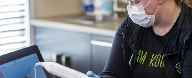 A Kohrs Orthodontics team member wearing a face mask and blue gloves uses a handheld 3D oral scanner to take digital impressions of a patient’s teeth while viewing the digital image on a nearby computer monitor in a modern dental office, representing how digital impressions transform your orthodontic experience