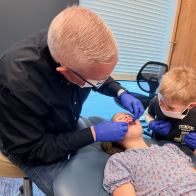 Orthodontist wearing gloves and a mask examining a young girl’s teeth in a dental chair while a young boy, also wearing a mask and gloves, observes in the treatment room, showing the right time for orthodontic treatment, representing right age for orthodontic intervention