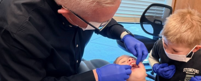 Orthodontist wearing gloves and a mask examining a young girl’s teeth in a dental chair while a young boy, also wearing a mask and gloves, observes in the treatment room, showing the right time for orthodontic treatment, representing right age for orthodontic intervention